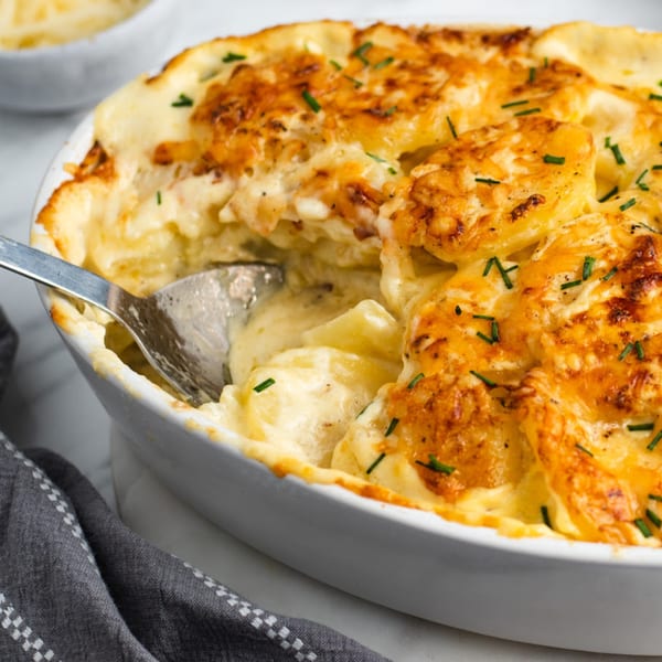Close-up of golden-brown scalloped potatoes with chives in a white baking dish, with a spoon.