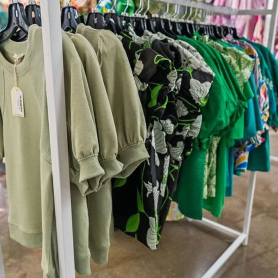 A clothing rack displays various green and patterned tops, some with price tags, in a retail setting.