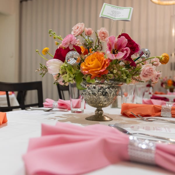 A vibrant floral centerpiece with disco ball accents on a white table set with pink and orange napkins.