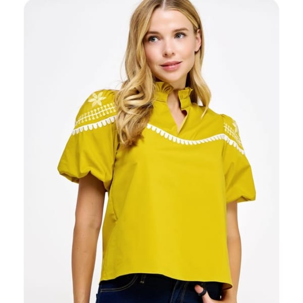 Blonde woman in a mustard yellow blouse with white embroidered details and dark jeans.