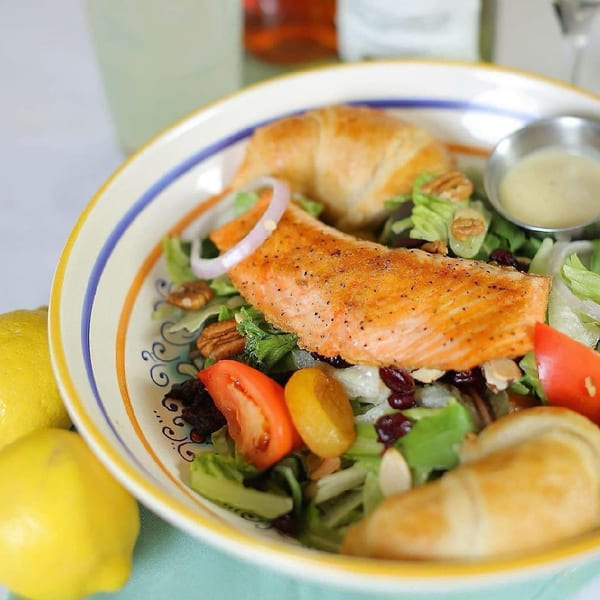 A salmon salad with greens, tomatoes, onions, nuts, and croissants in a colorful bowl.
